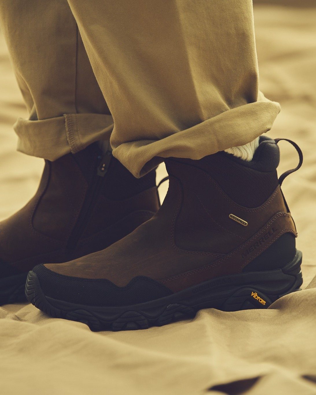 Brown waterproof winter boots with black soles worn with tan trousers on sand.