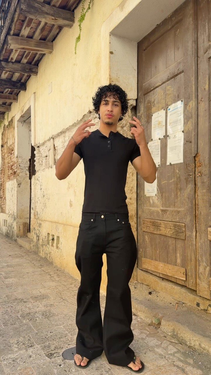 Young man wearing a black polo shirt and wide-leg black trousers outdoors.