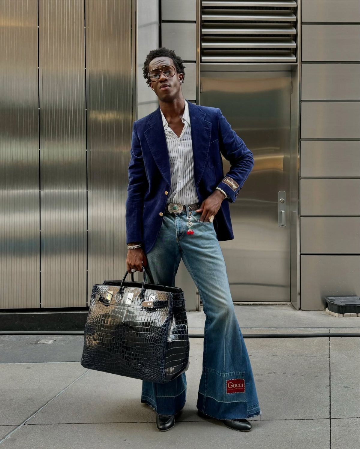 Black man in a blue blazer and flared jeans holding a large black bag.