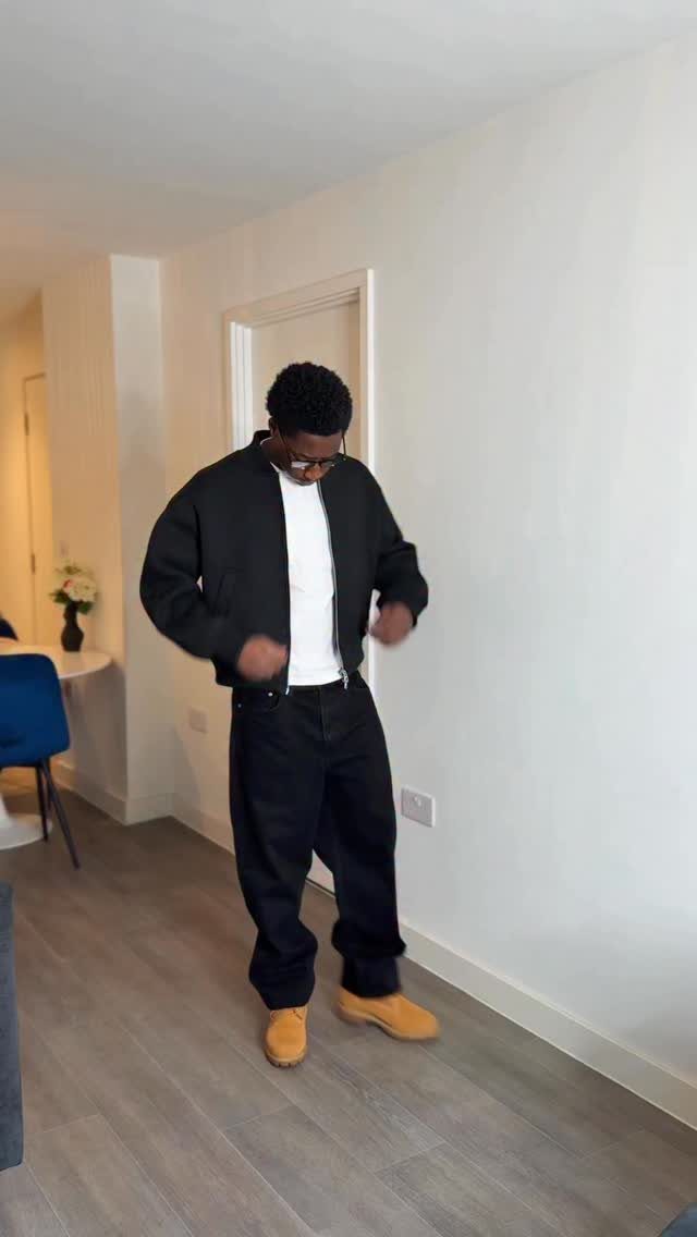 Black man wearing a black bomber jacket, white t-shirt, black trousers, and tan boots.