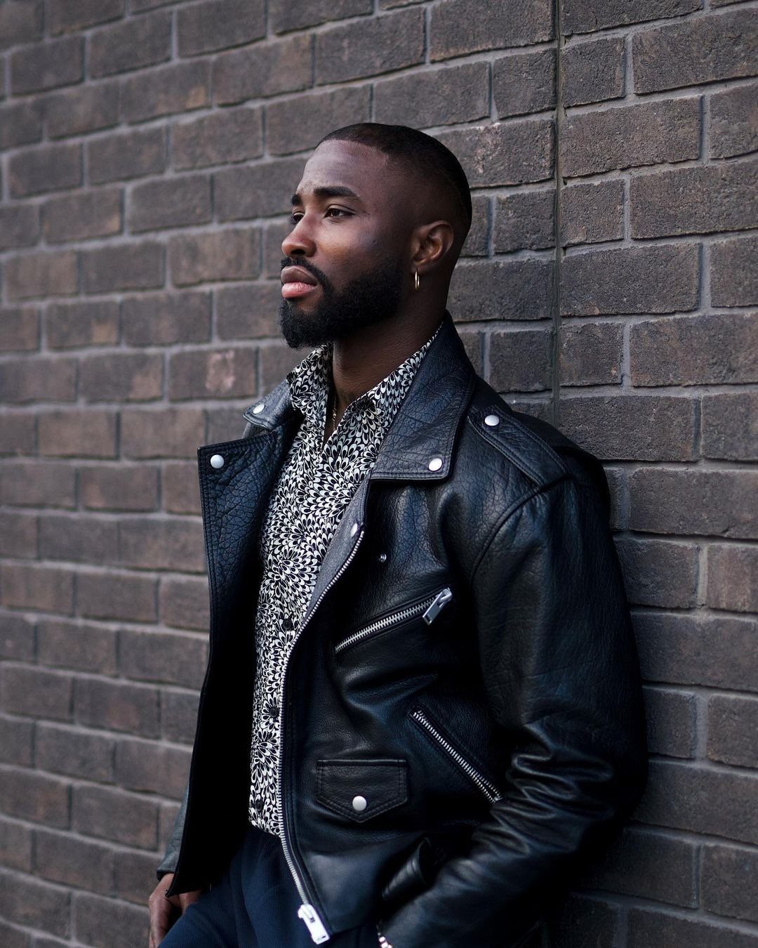 Black man wearing a black leather jacket and patterned shirt against a brick wall