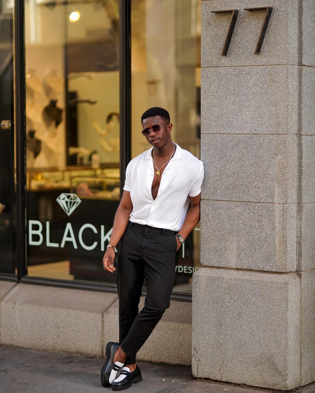 Black man wearing a white button-down shirt and black chinos leaning against a wall.