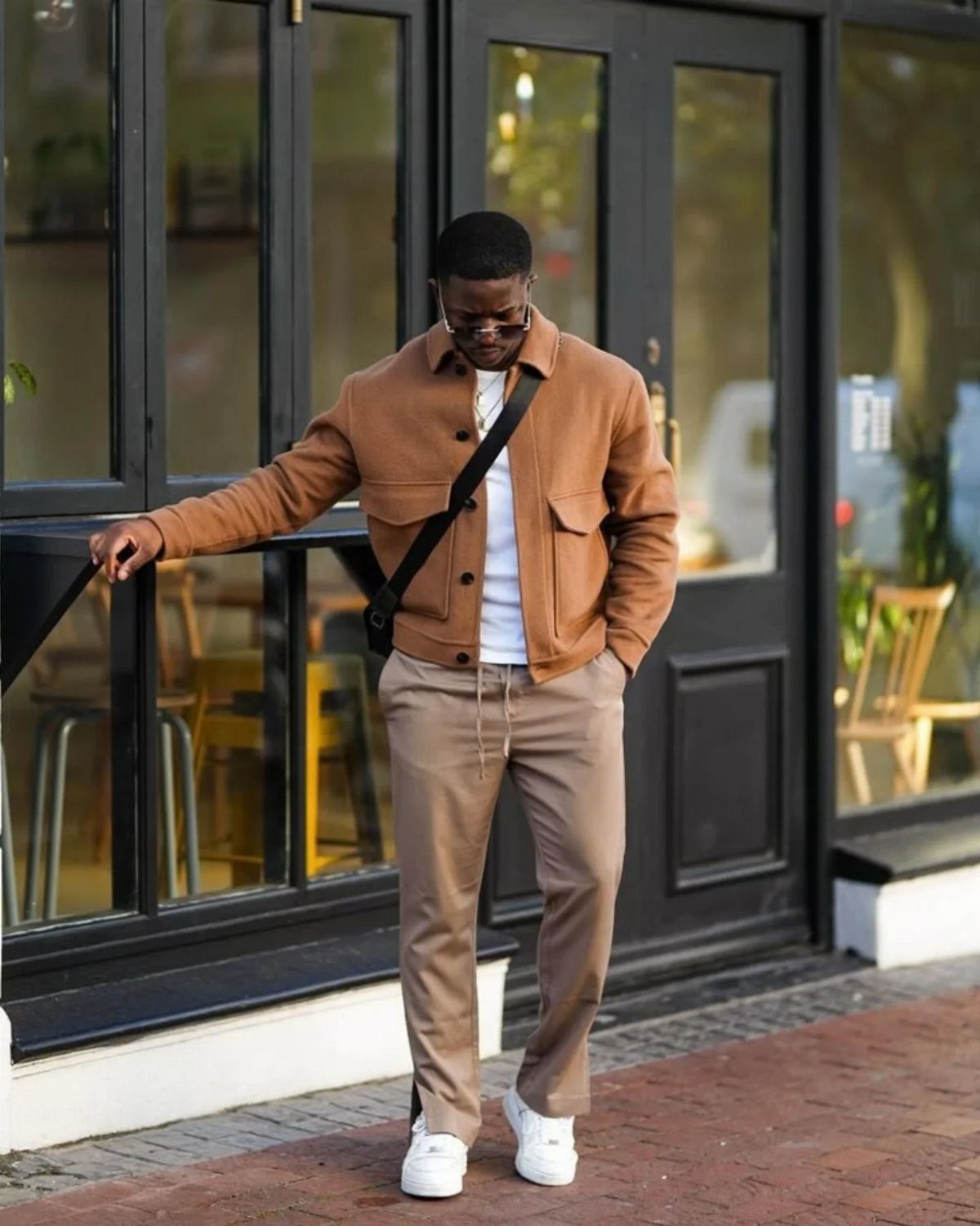 Black man in brown jacket and tan trousers posing by a storefront.