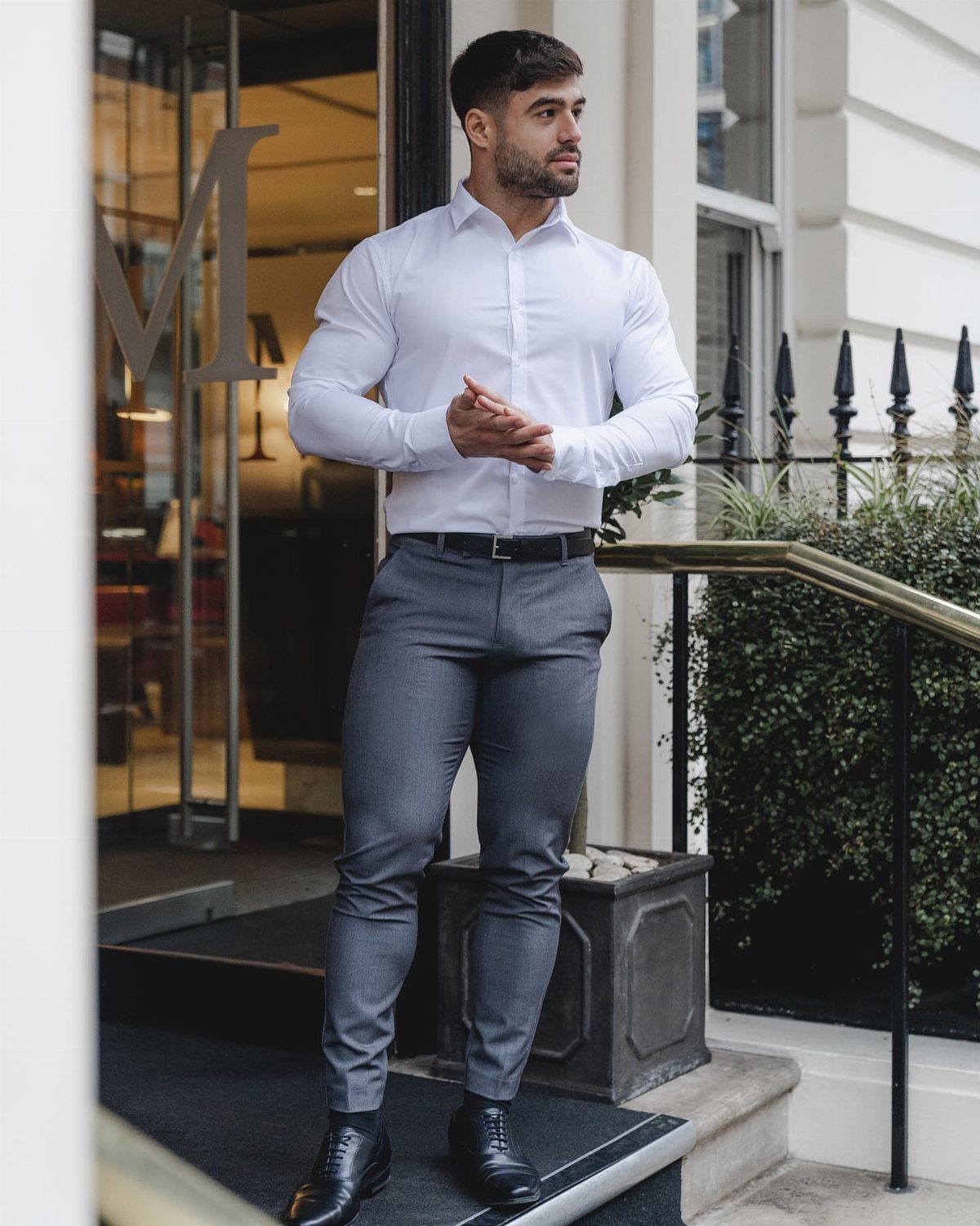 Man in white dress shirt and grey trousers posing outside a building.