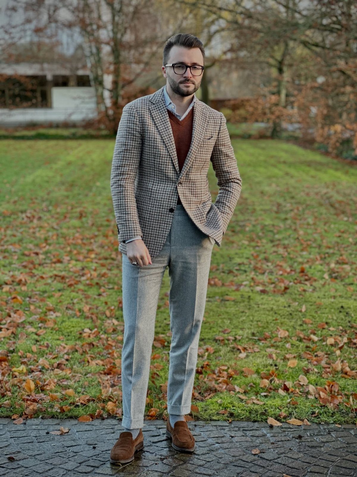 Man wearing a patterned blazer, burgundy sweater, and grey trousers in an autumn setting.