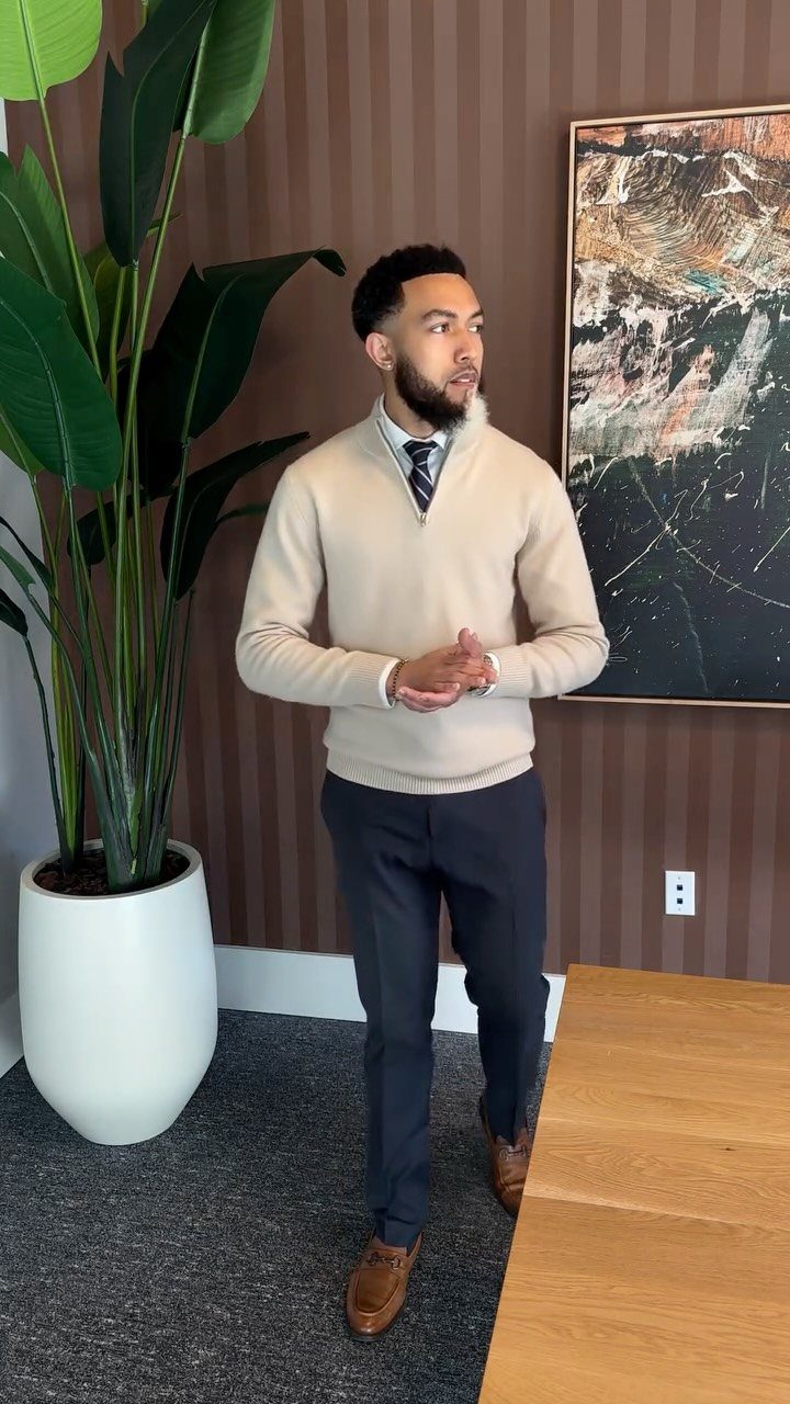 Man in beige sweater, tie, and navy trousers posing in a modern office.