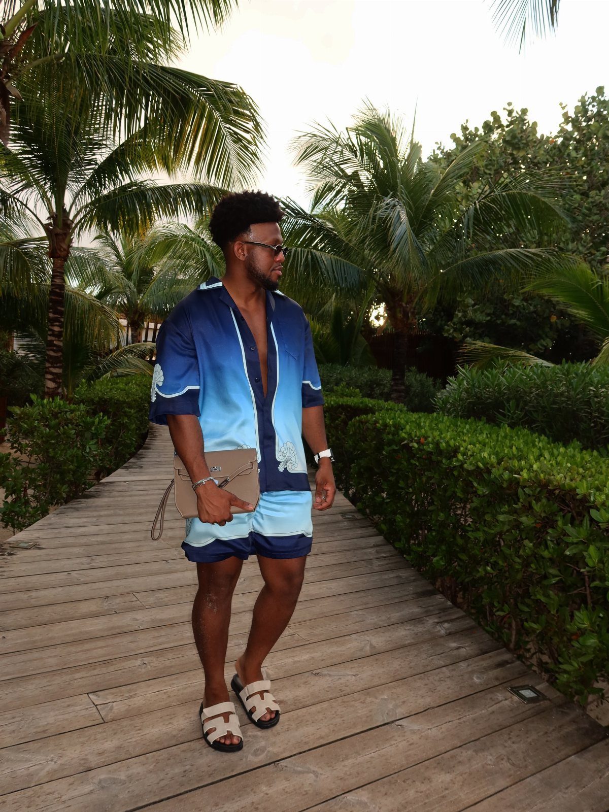 Man in blue gradient resort set and sandals standing on a tropical wooden boardwalk.