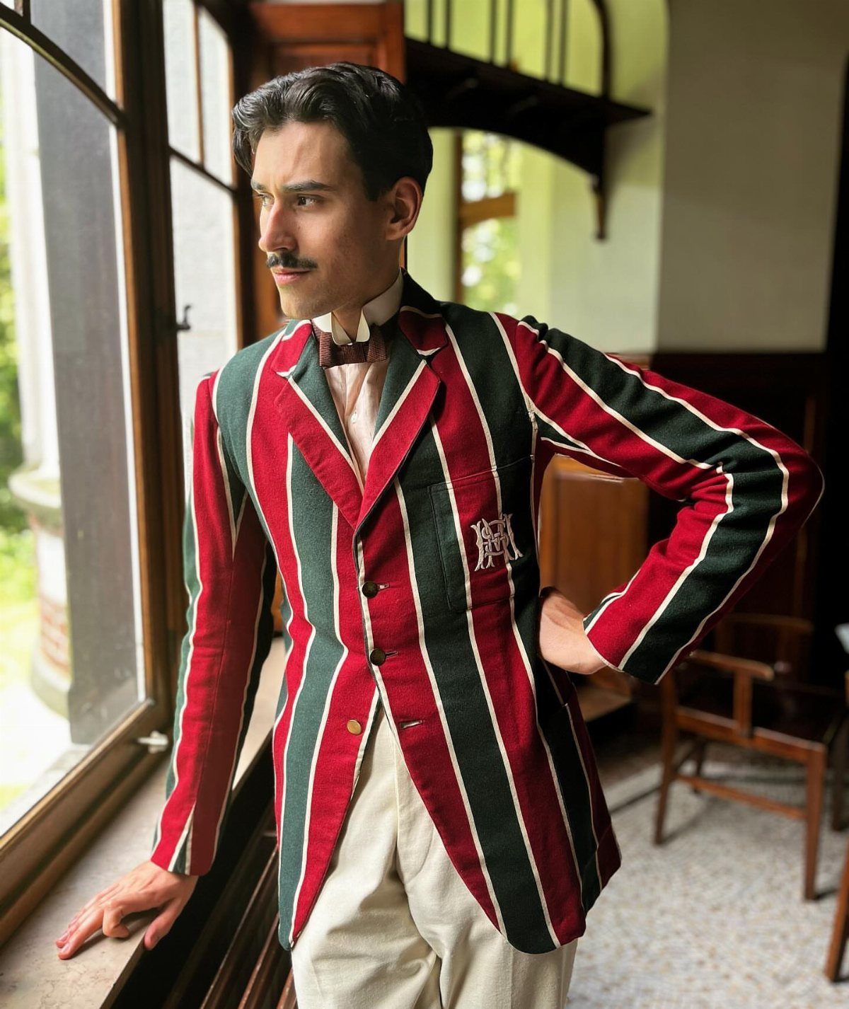 Man in red and green striped blazer and high collar standing by window.