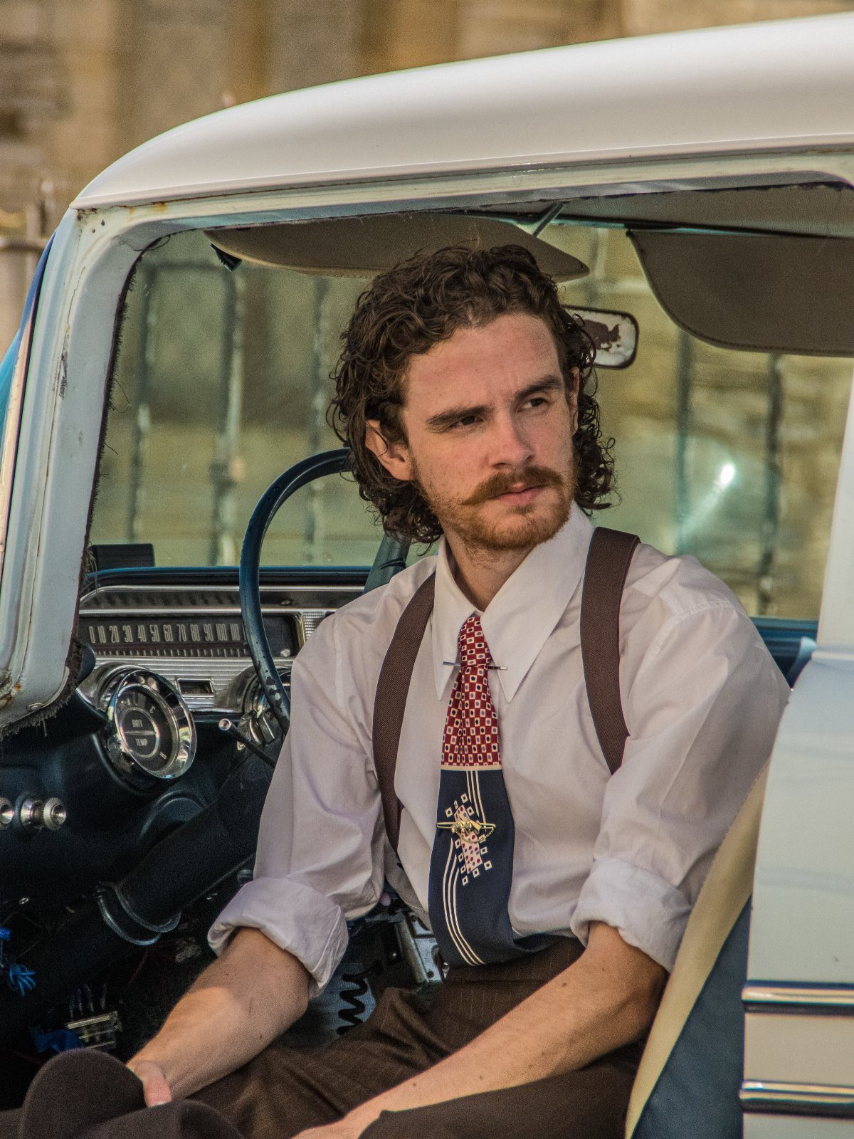 Man in vintage white shirt, suspenders, and tie sitting in a classic car.