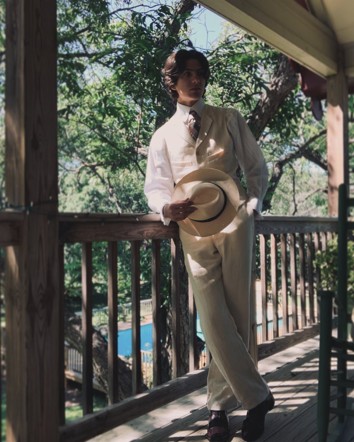 Man in a cream-colored summer suit holding a straw boater hat on a porch.