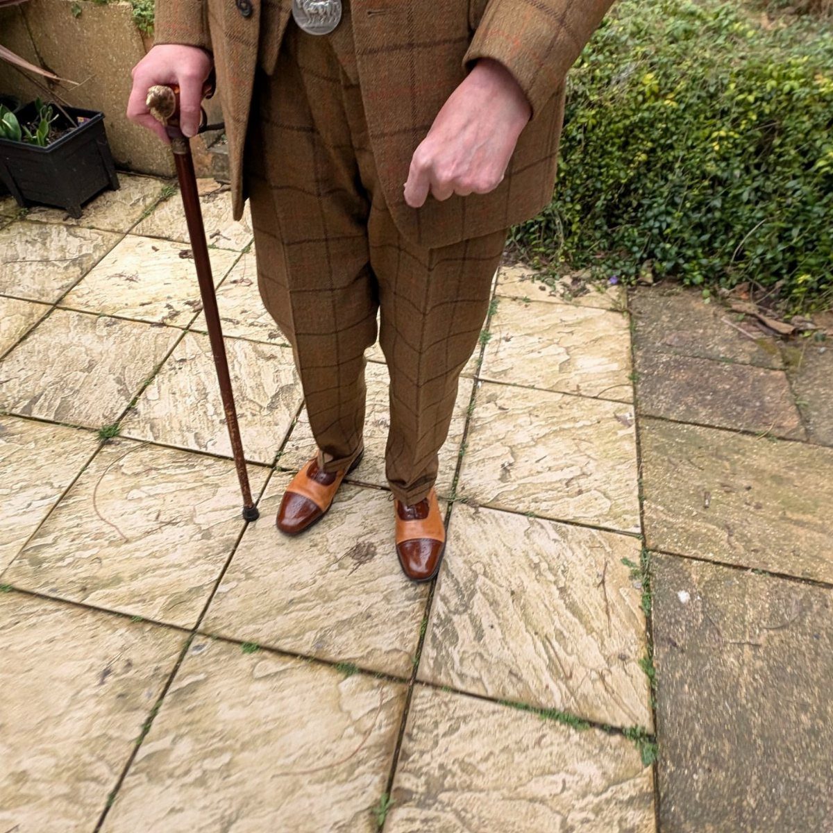Man in brown plaid suit and leather shoes holding a wooden walking cane.