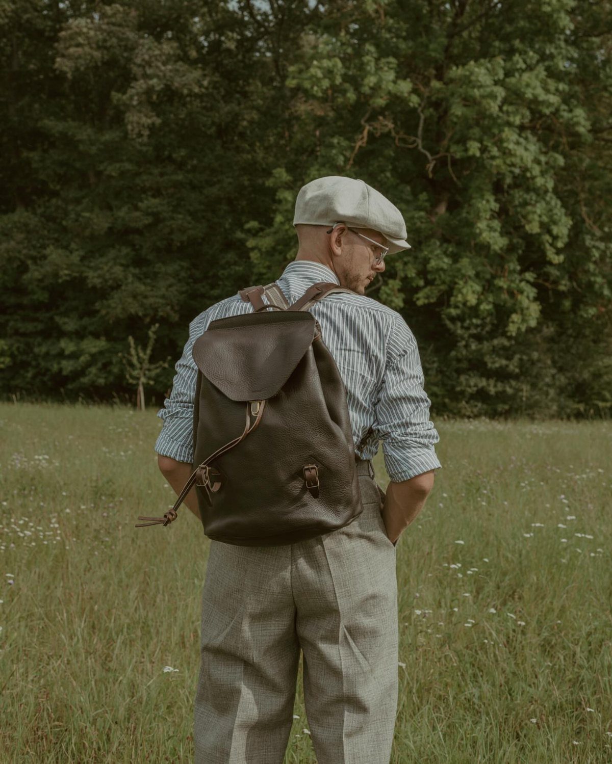Man in a flat cap and striped shirt wearing a leather backpack in a field.