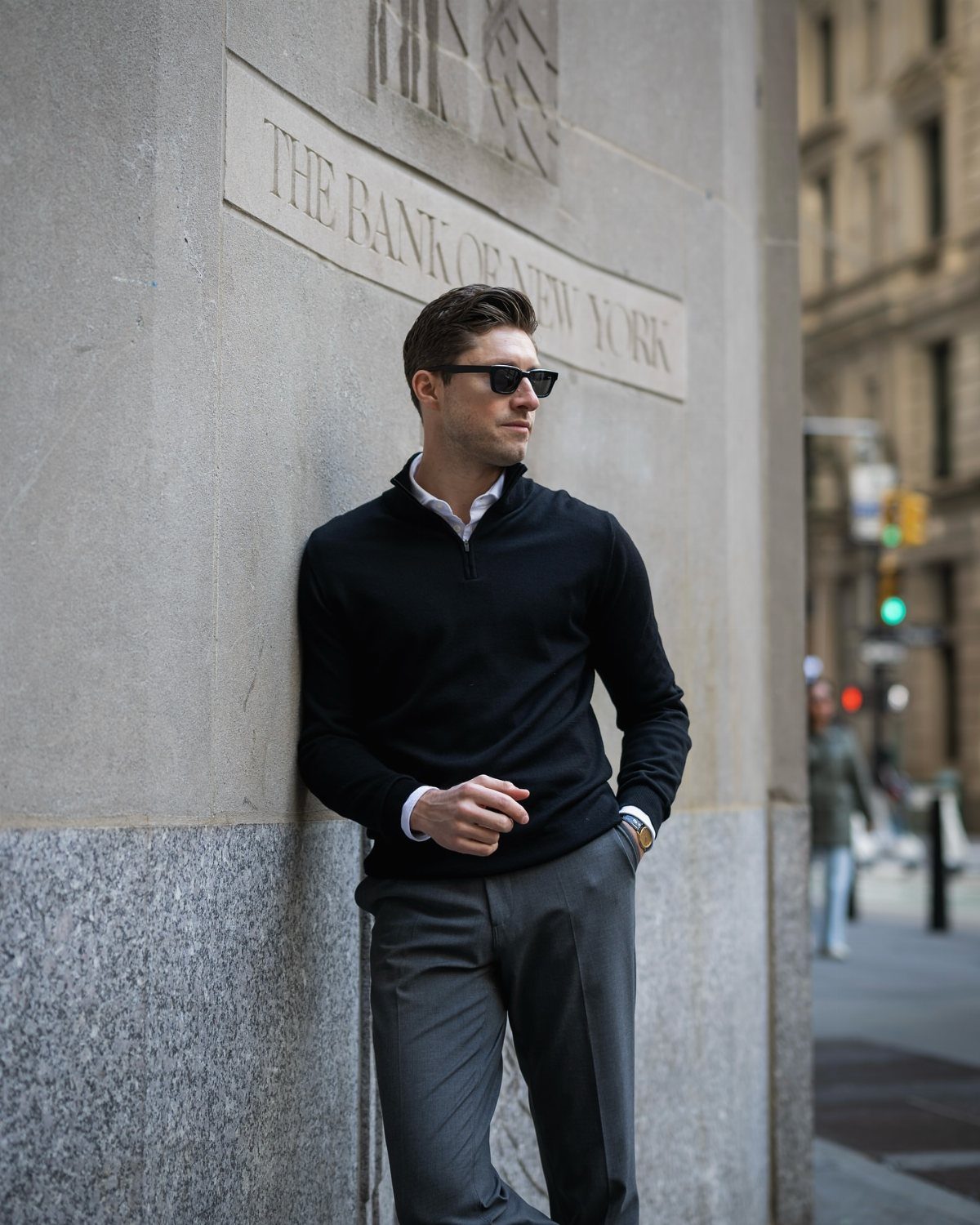 Casual Chic Dress Code Men: 16 Looks That Work in 2026 6 Man in black quarter-zip sweater and grey trousers leaning against a stone building wall.