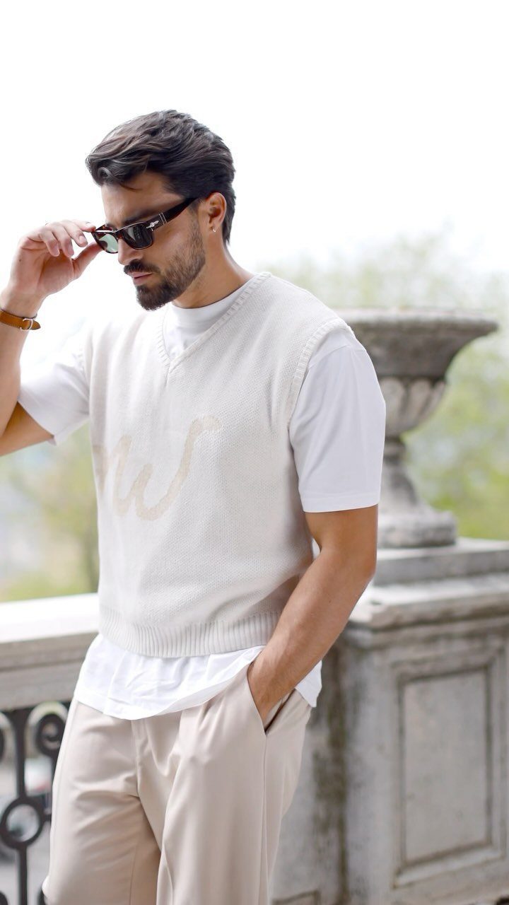 Casual Chic Dress Code Men: 16 Looks That Work in 2026 9 Man in a cream knit vest, white t-shirt, and beige trousers posing outdoors.