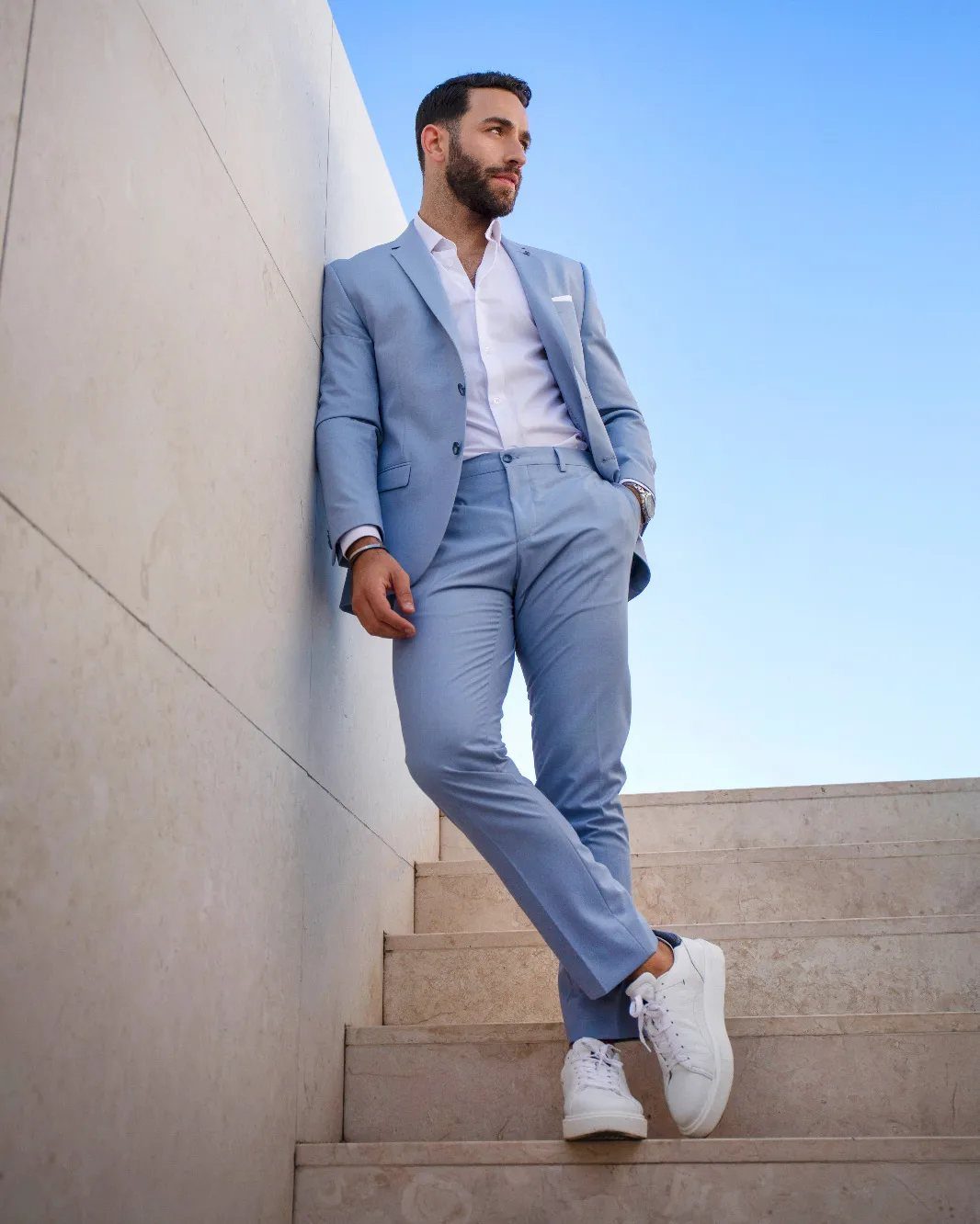 Man in a light blue suit and white sneakers leaning against a white wall.