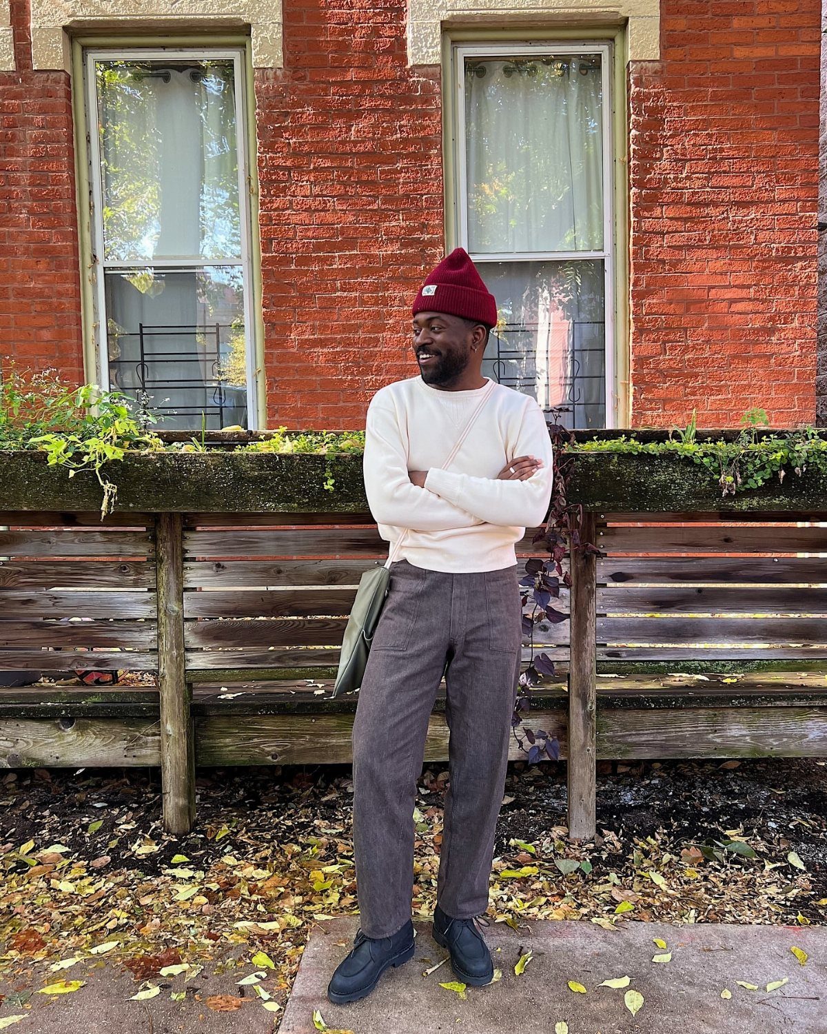 Man in red beanie and cream sweater posing against a red brick wall.