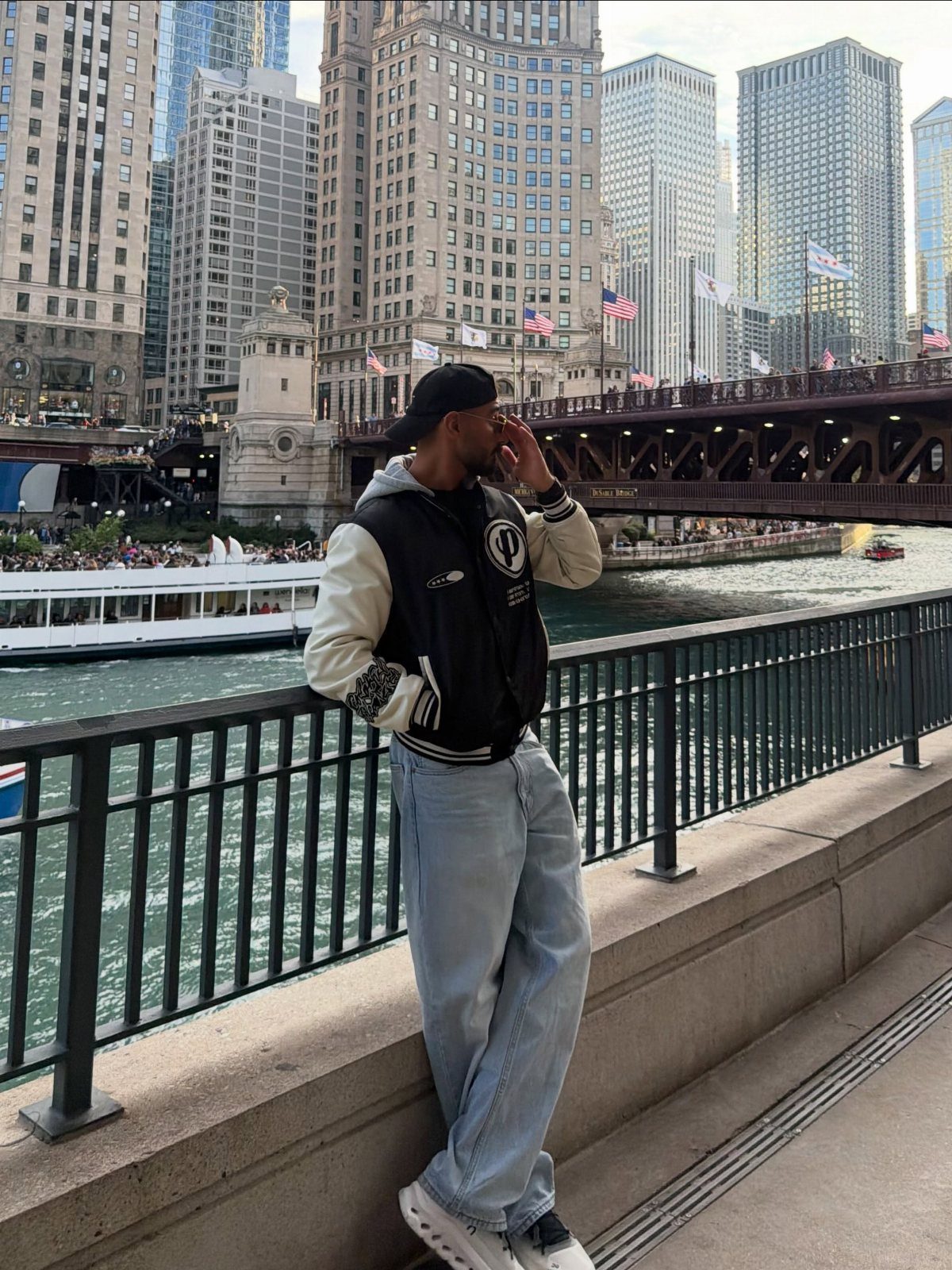 Man in varsity jacket and baseball cap posing by a Chicago riverwalk.