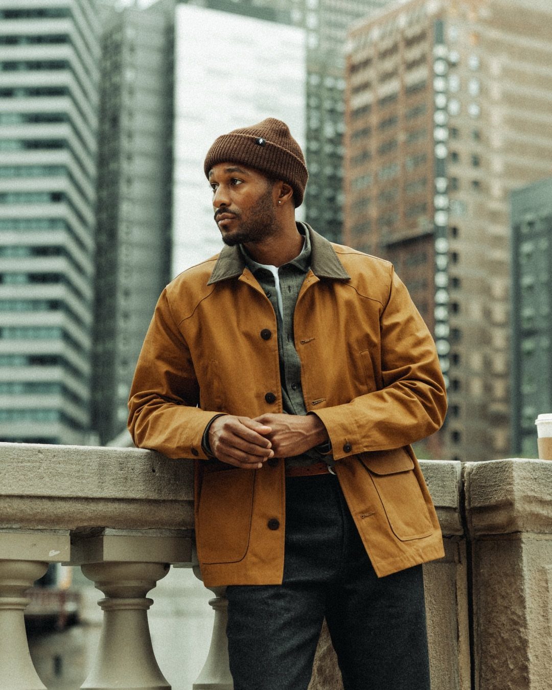 Man in mustard yellow jacket and brown beanie posing in a city setting.