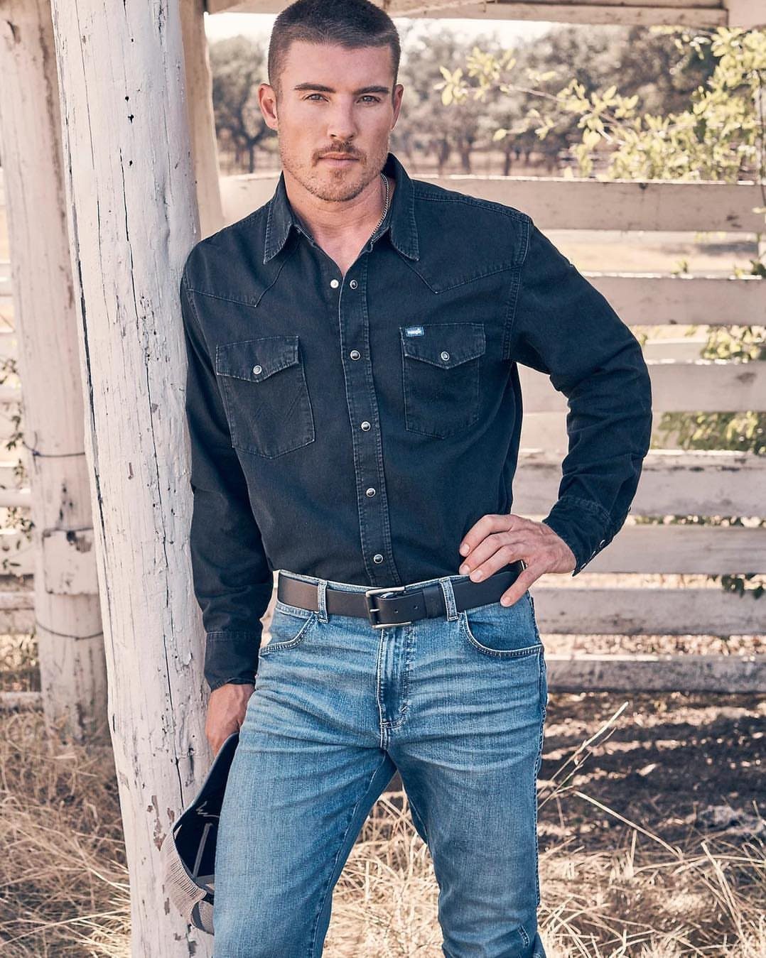Man wearing a black button-down shirt and blue denim jeans leaning on a post.
