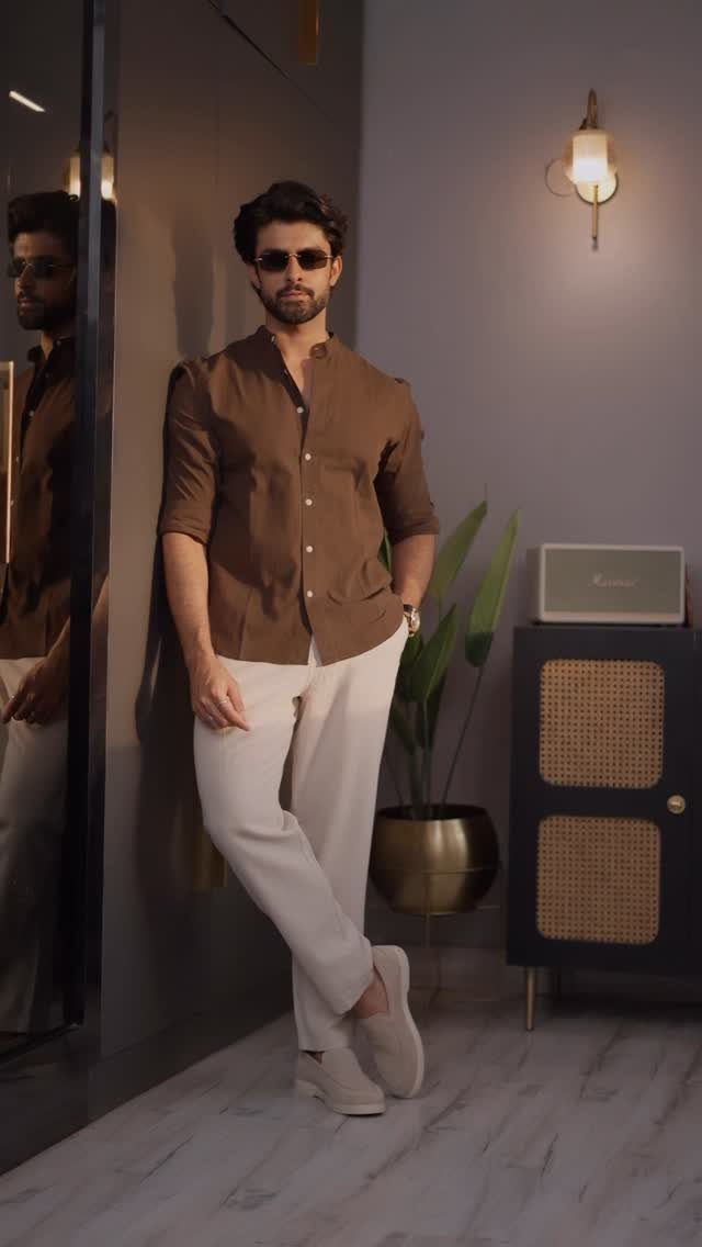 Man in brown short-sleeved shirt and off-white trousers leaning against a wall.