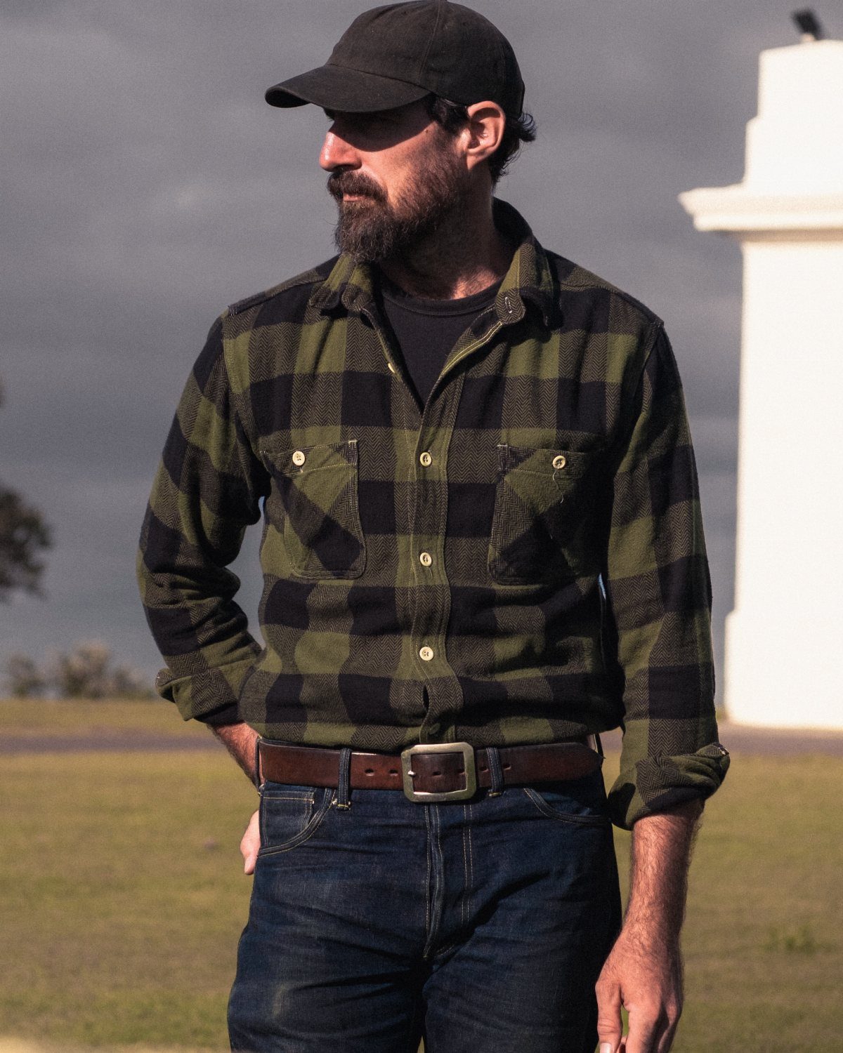 Bearded man wearing a green and black checkered flannel shirt and dark denim jeans.