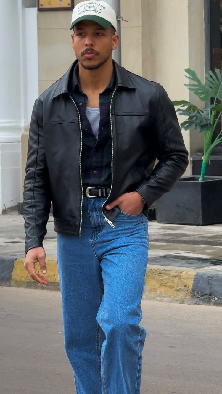 Man wearing a black leather jacket, navy shirt, and blue jeans walking outdoors.