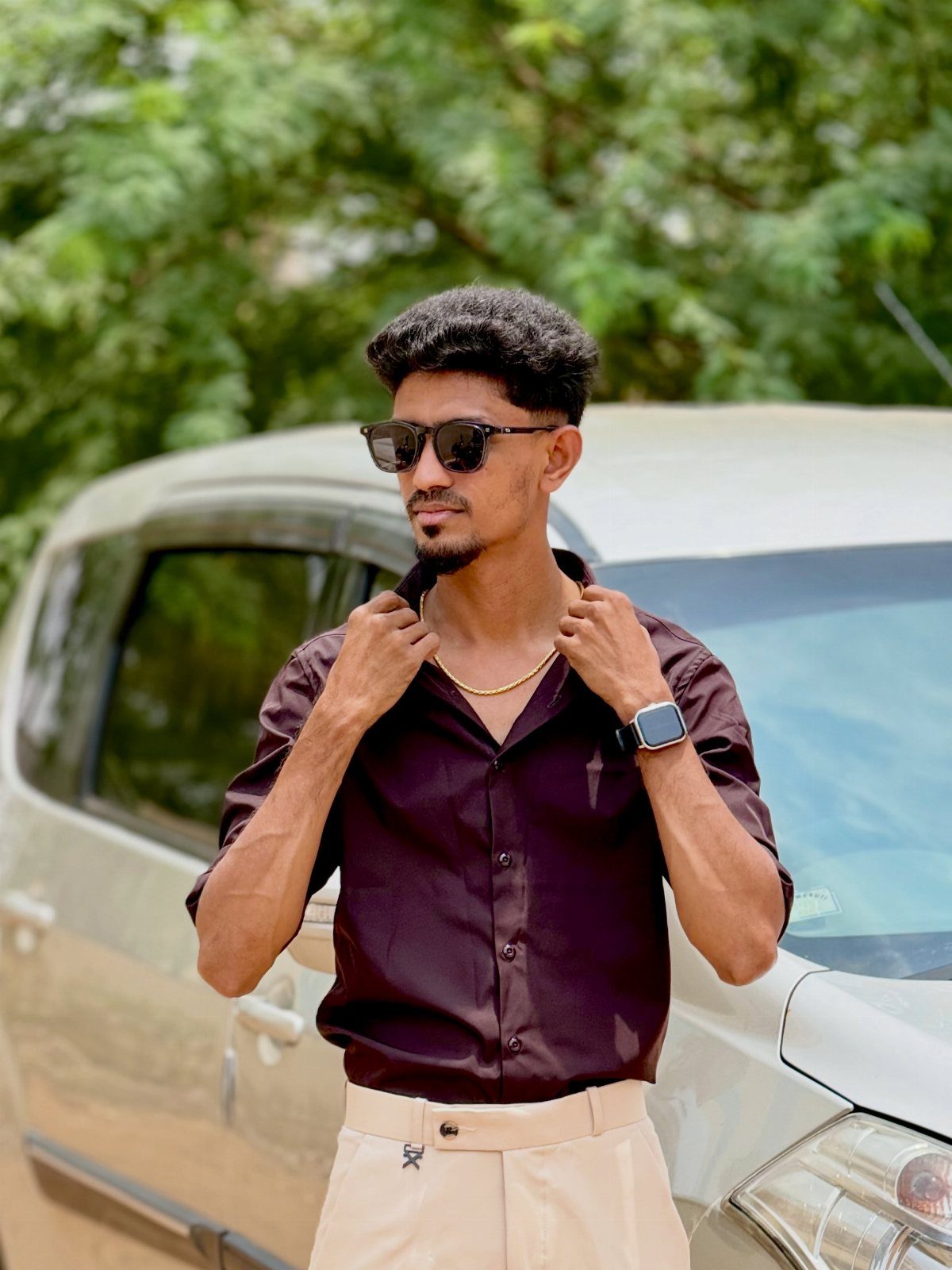 Man in maroon button-down shirt and beige trousers posing by a silver car.