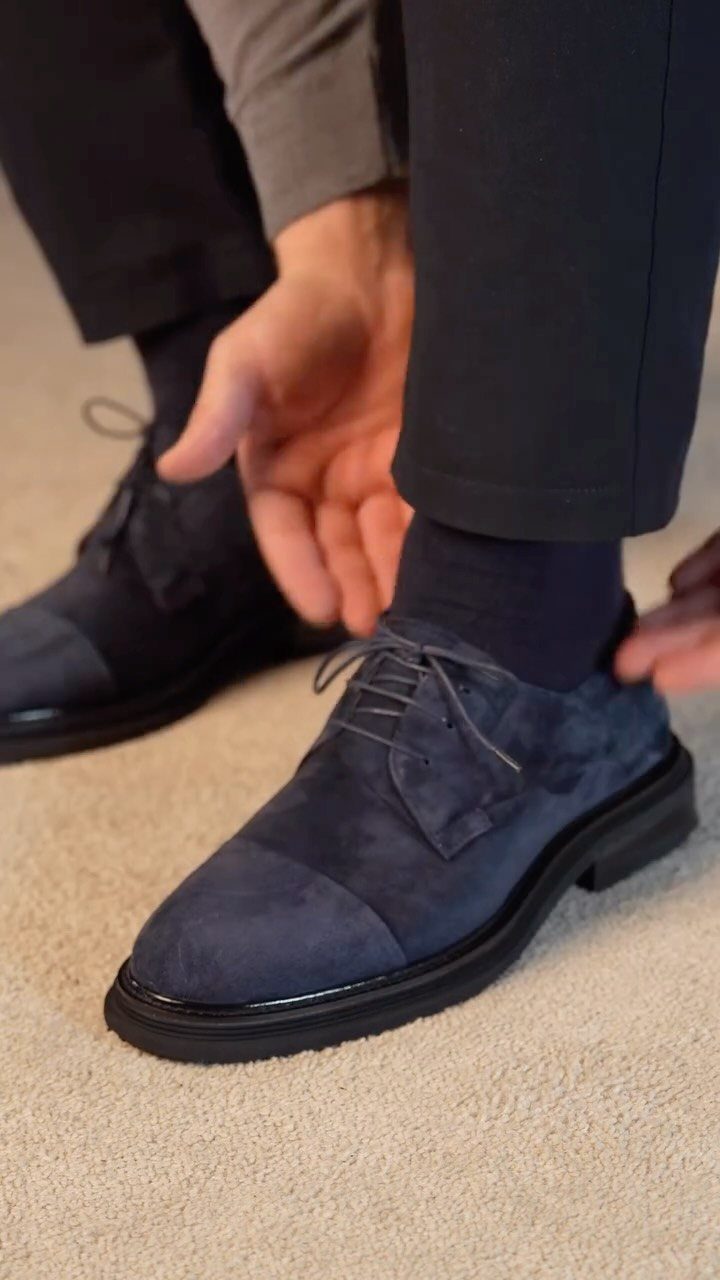 Navy blue suede derby shoes worn with dark trousers on a beige carpet.