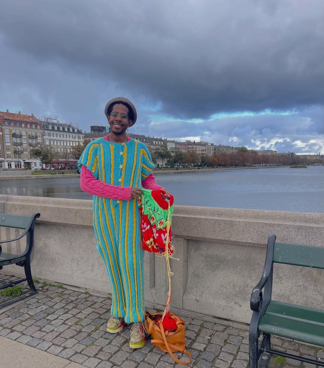 Man in vibrant striped oversized clothing and pink shirt posing by a city lake.