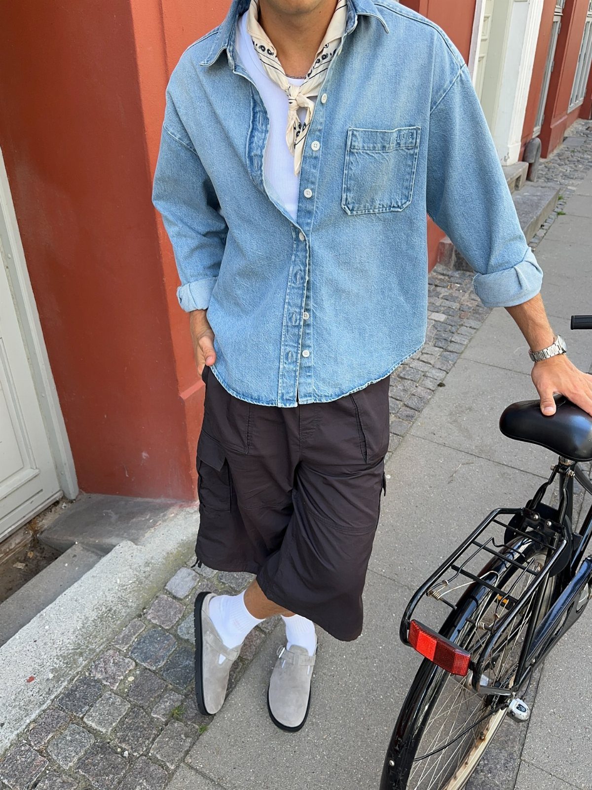 Man in oversized denim shirt and black cropped trousers standing by a bicycle.