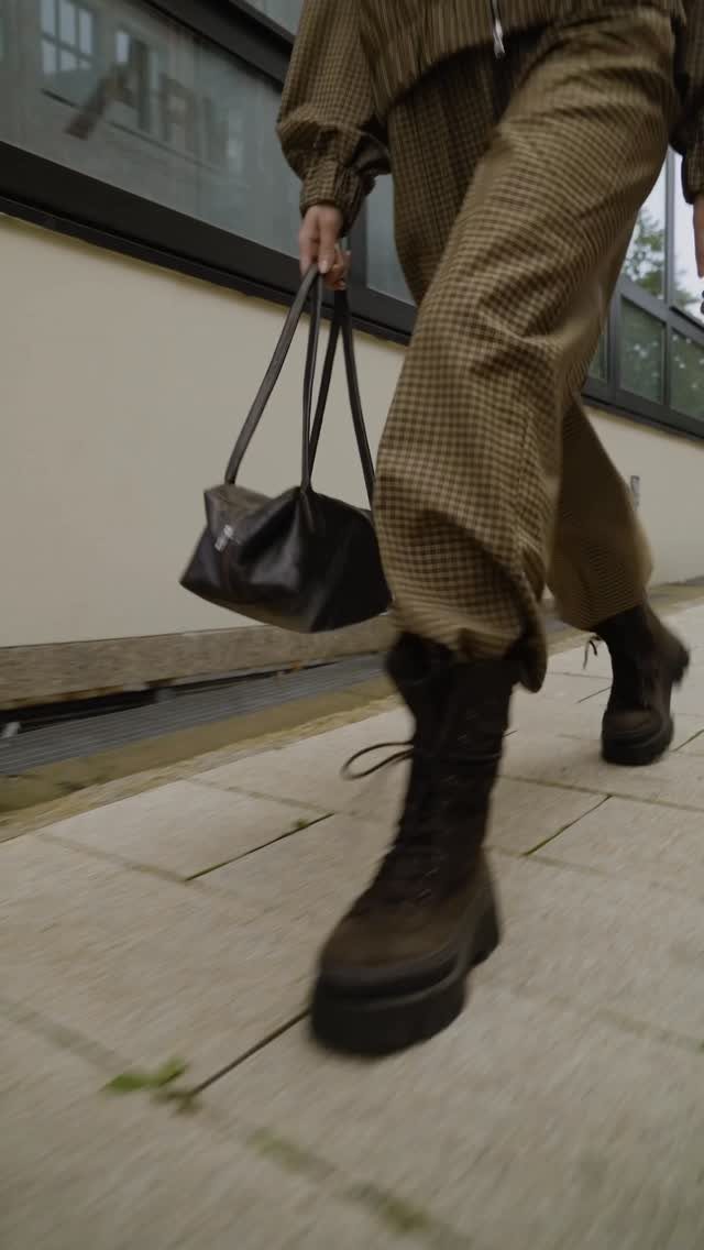 Chunky black lace-up boots and brown checkered trousers on a person walking outdoors.