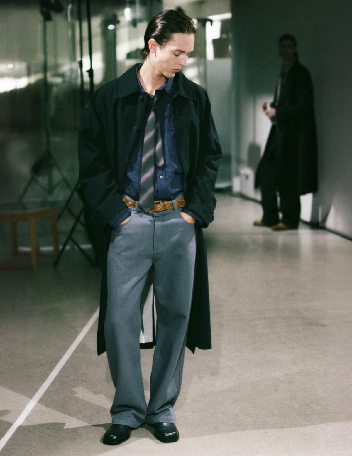 Man in a long black coat and grey wide-leg trousers in a minimalist setting.