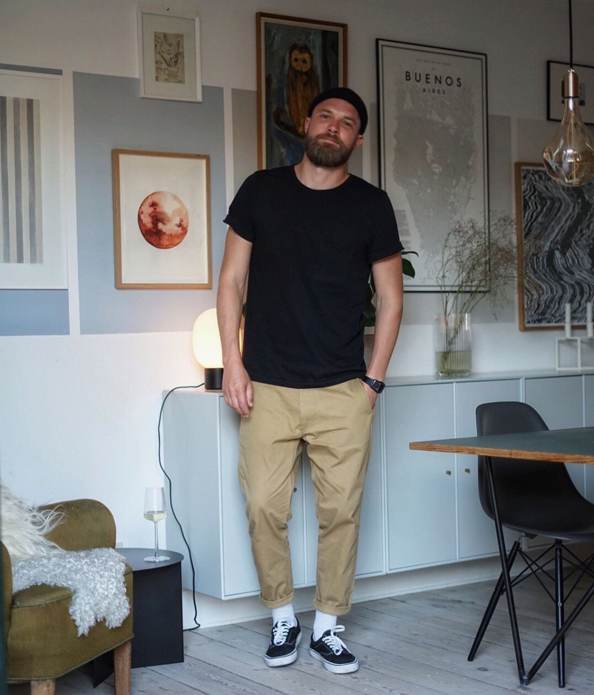 Man in black t-shirt and tan trousers posing in a modern apartment.