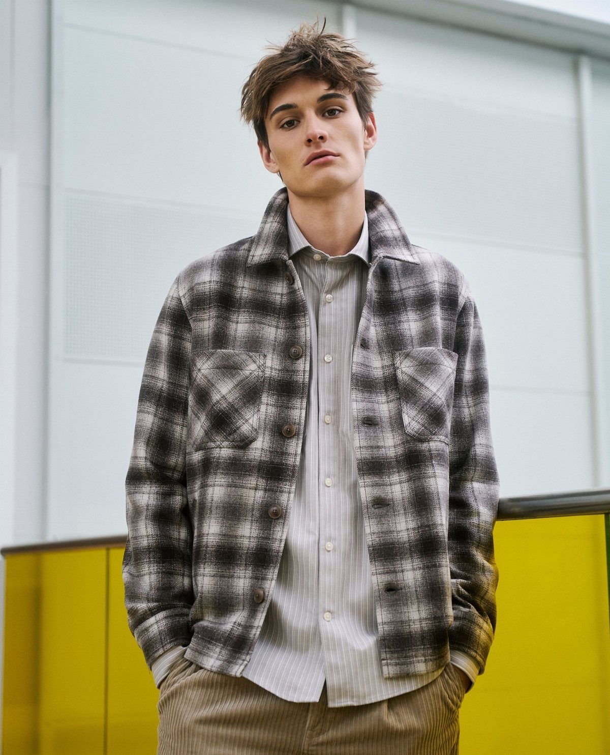 Man wearing an oversized plaid jacket and striped shirt in a modern urban setting.