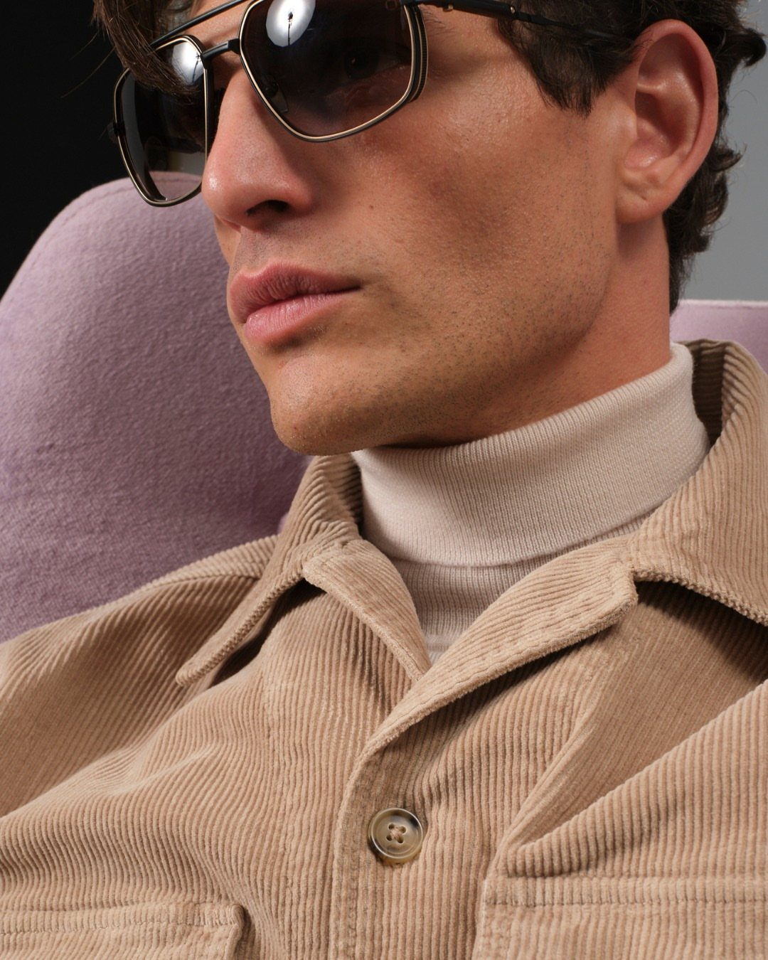 Man wearing dark sunglasses, white turtleneck, and tan corduroy jacket against purple background.