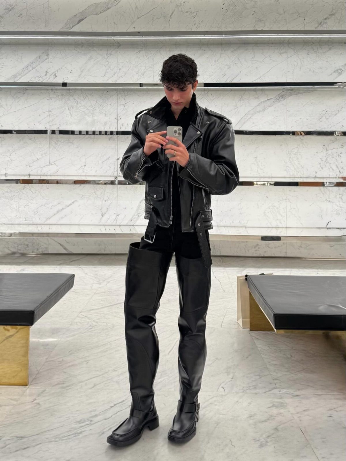 Man in black leather biker jacket and thigh-high leather boots taking a mirror selfie.