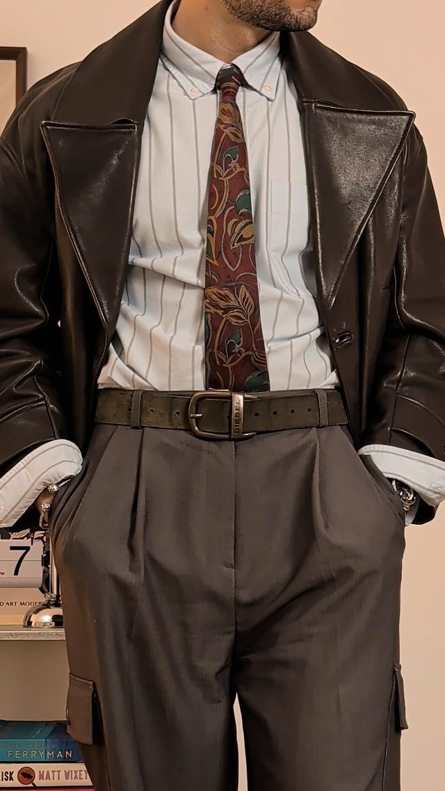 Man in oversized black leather jacket, pinstriped shirt, patterned tie, and pleated grey trousers.