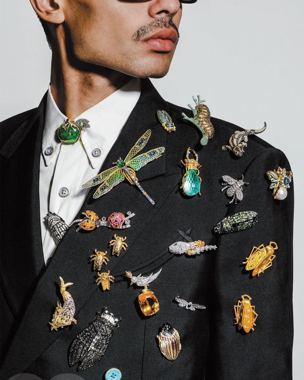 Man in black suit jacket decorated with many oversized jeweled insect brooches.