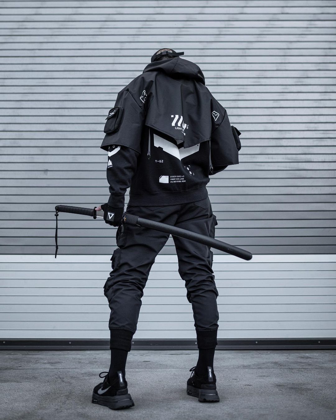 Man in black techwear outfit holding a sword against a grey ribbed wall