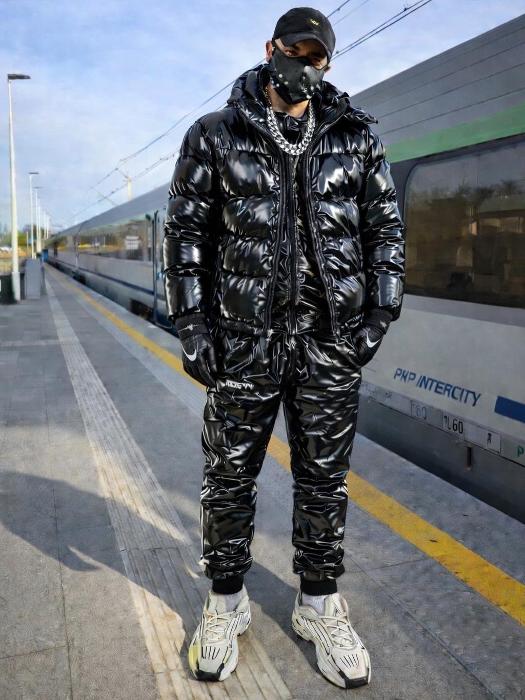 Man in glossy black puffer outfit and face mask standing by a train.