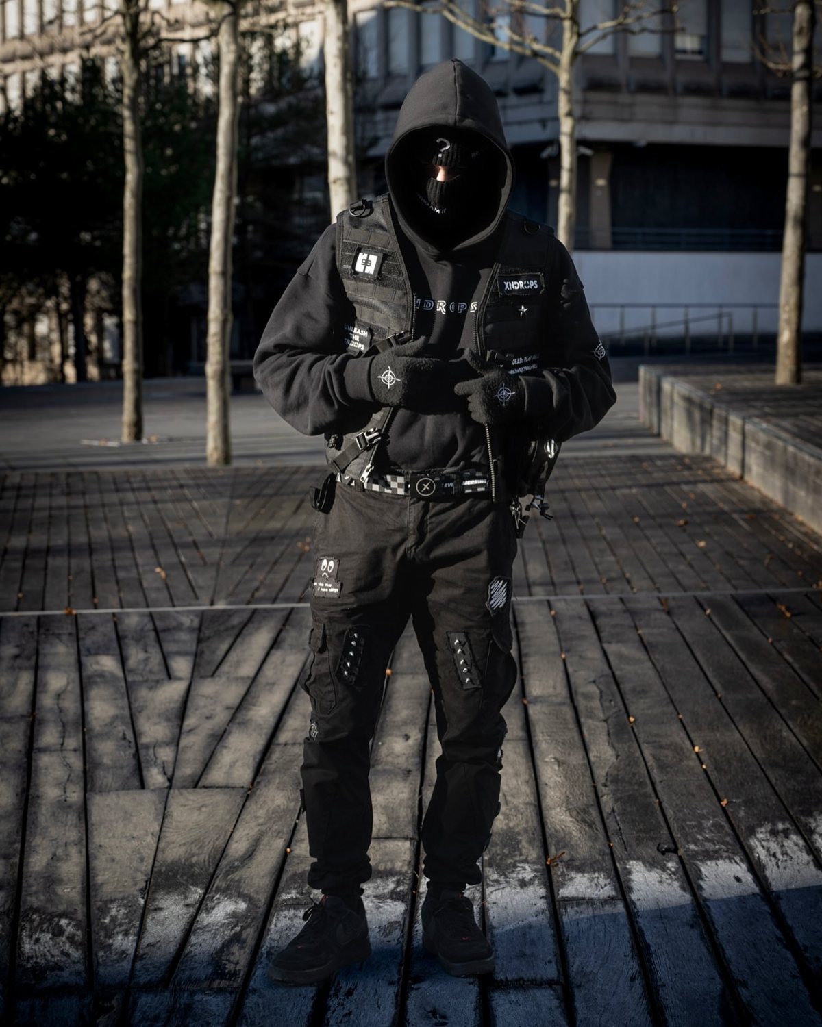 Man in black cyberpunk tactical gear with layered hoodie and vest on wooden boardwalk