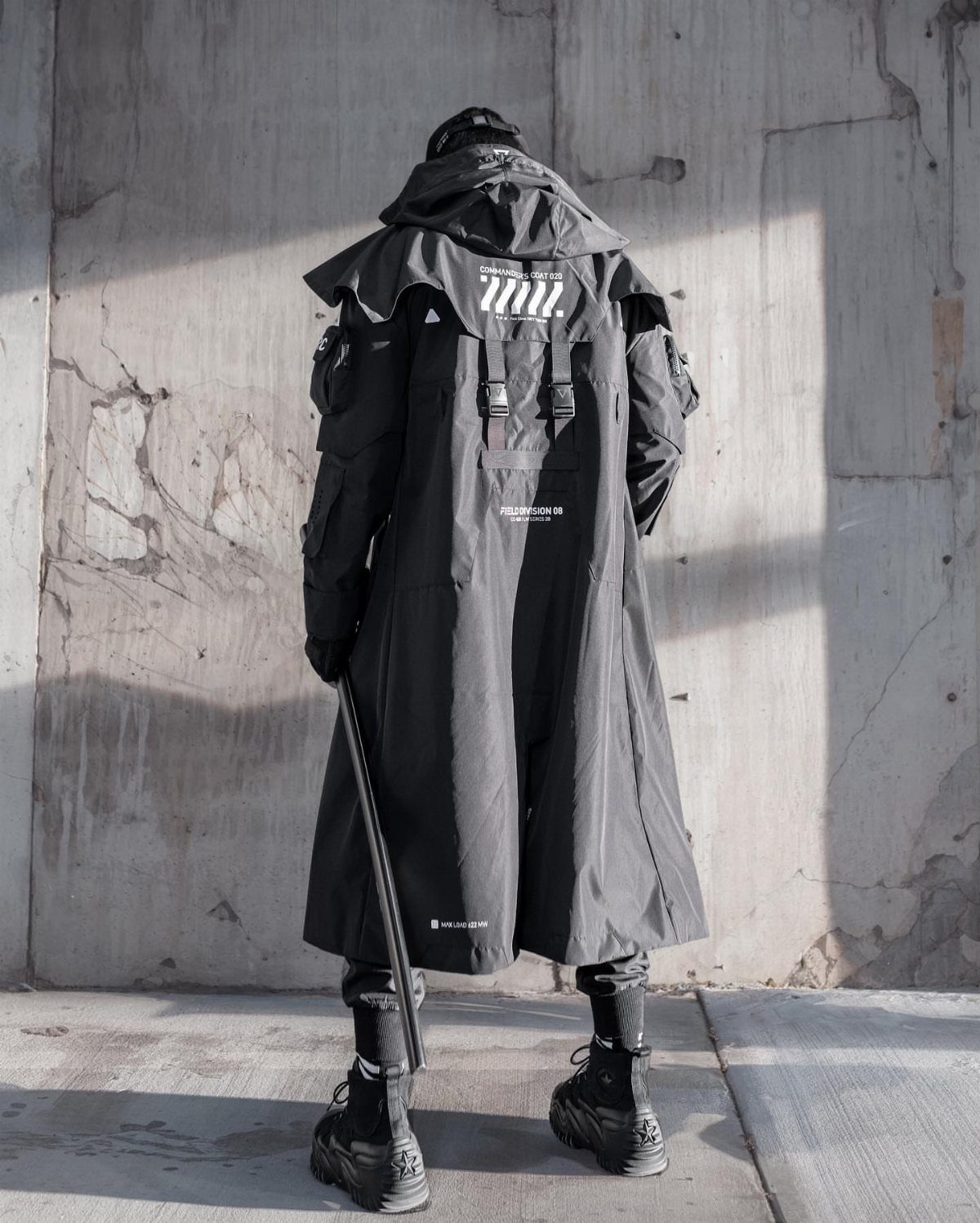 Man in a long black technical cyberpunk coat and chunky boots against concrete.