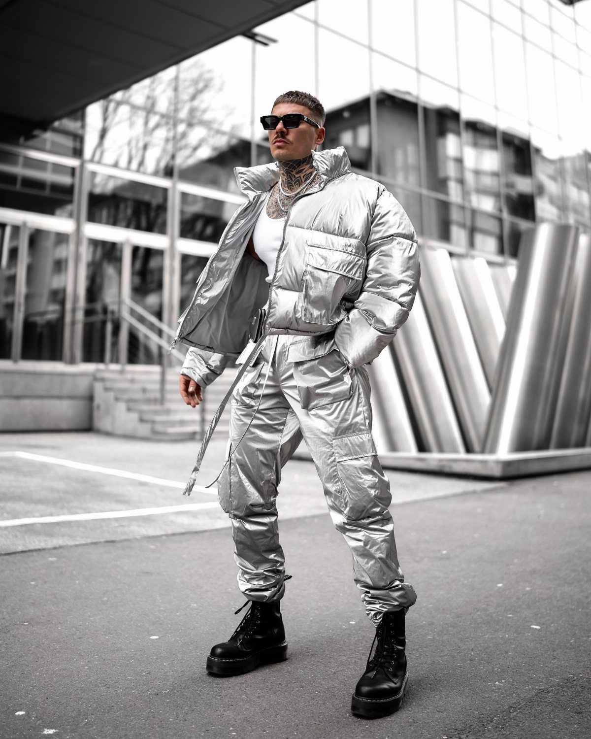 Man in a silver metallic puffer jacket and matching trousers standing by a building.