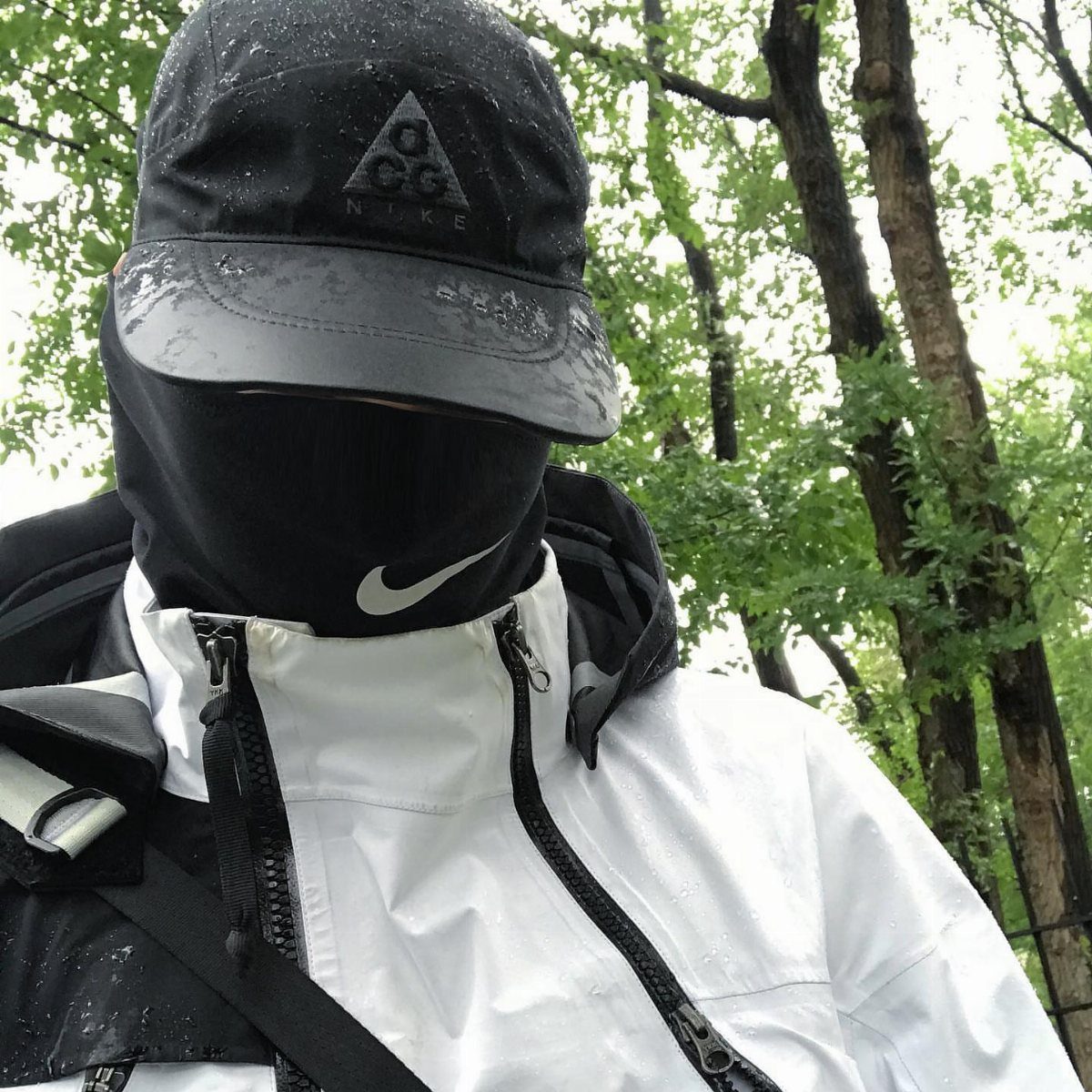 Person in a white techwear rain shell, black cap, and face mask outdoors.