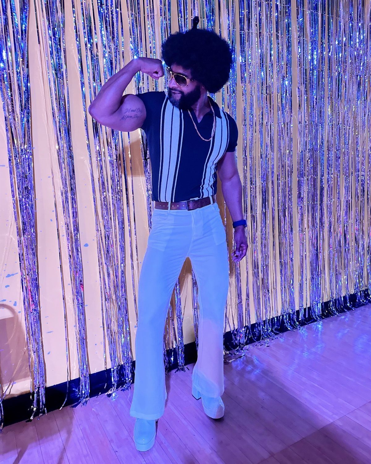 Man with afro wearing striped shirt and white bell-bottoms against silver tinsel backdrop.