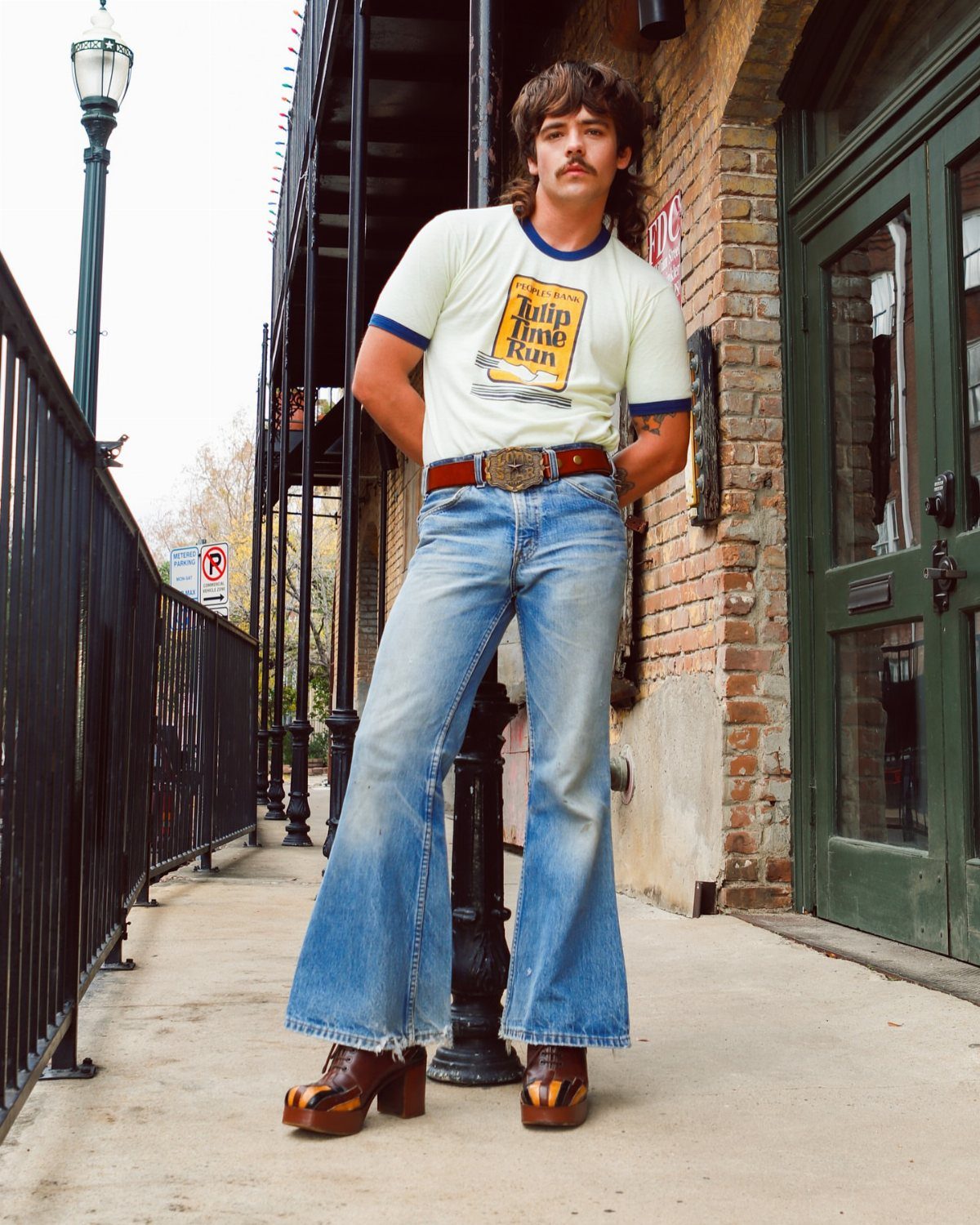 Man in flared jeans and brown platform boots posing on a city sidewalk.