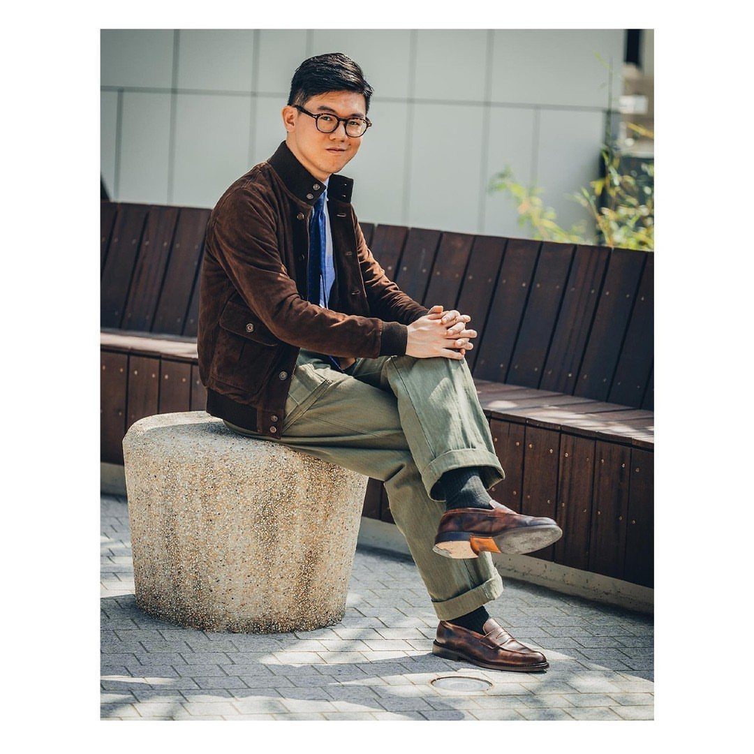 Man in brown suede jacket and olive trousers sitting outdoors on concrete bollard