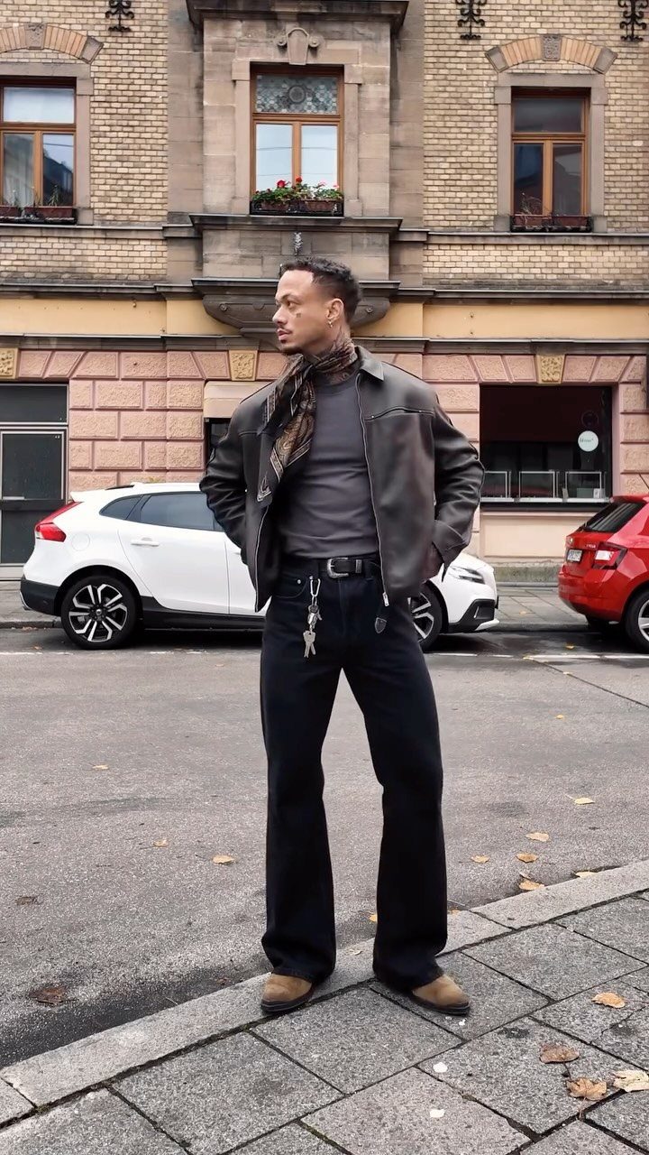 Man in leather jacket and black flared trousers standing on a city street.