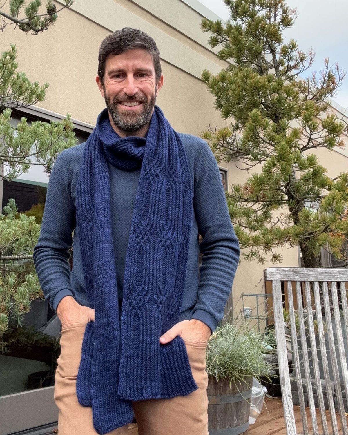 Man wearing a long blue knitted scarf, blue sweater, and tan trousers outdoors.
