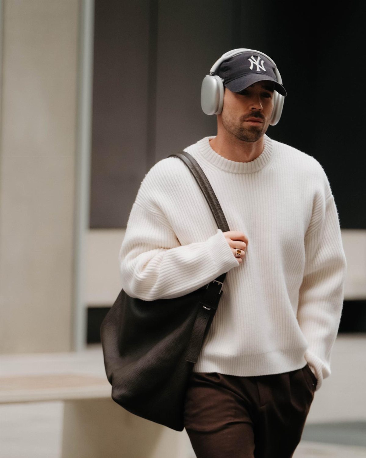 Man in white oversized knit sweater, brown trousers, and navy baseball cap.